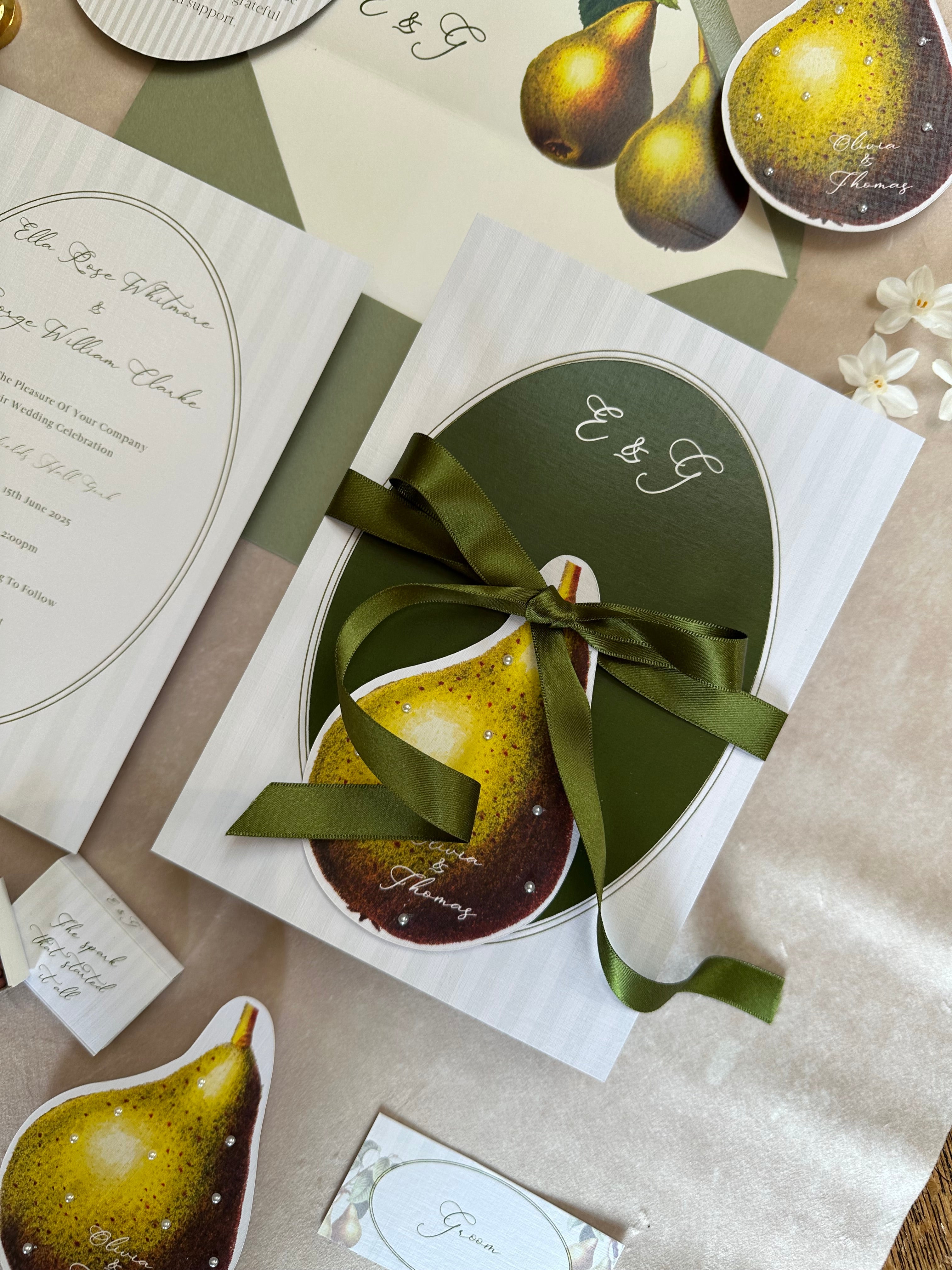 Close-up of layered wedding stationery set with satin ribbon, botanical pears, guest name tag, matchbox favour, and elegant invitation card